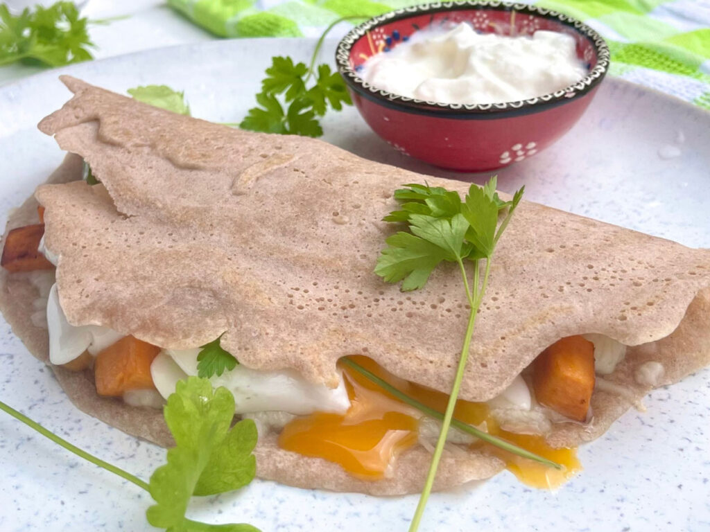 Eine herzhafte Galette aus Buchweizenmehl, gefüllt mit Süßkartoffeln, Ei und Käse, garniert mit frischer Petersilie, auf einem weißen Teller.