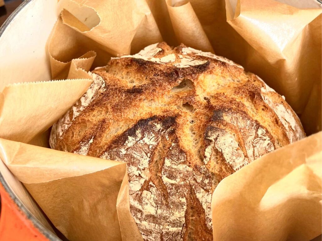 Brot selber backen, Ein frisch gebackenes Brot in einem gusseisernen Topf, umgeben von Backpapier.