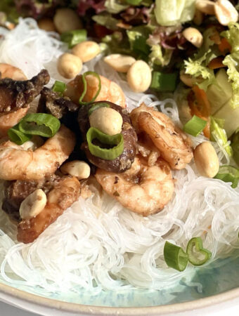 Nahaufnahme Reisnudelsalat mit gebratenen Garnelen, Pilzen und Erdnüssen. Der Salat enthält grüne Blätter, Frühlingszwiebeln und ist auf einem Teller angerichtet