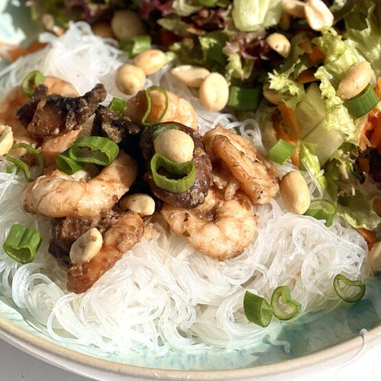 Nahaufnahme Reisnudelsalat mit gebratenen Garnelen, Pilzen und Erdnüssen. Der Salat enthält grüne Blätter, Frühlingszwiebeln und ist auf einem Teller angerichtet