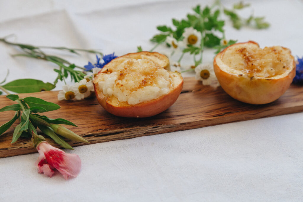 Zwei gebackene Apfelhälften, gefüllt mit Milchreis und karamellisierter Oberfläche, dekorativ auf einem Holzbrett mit Blumen arrangiert.