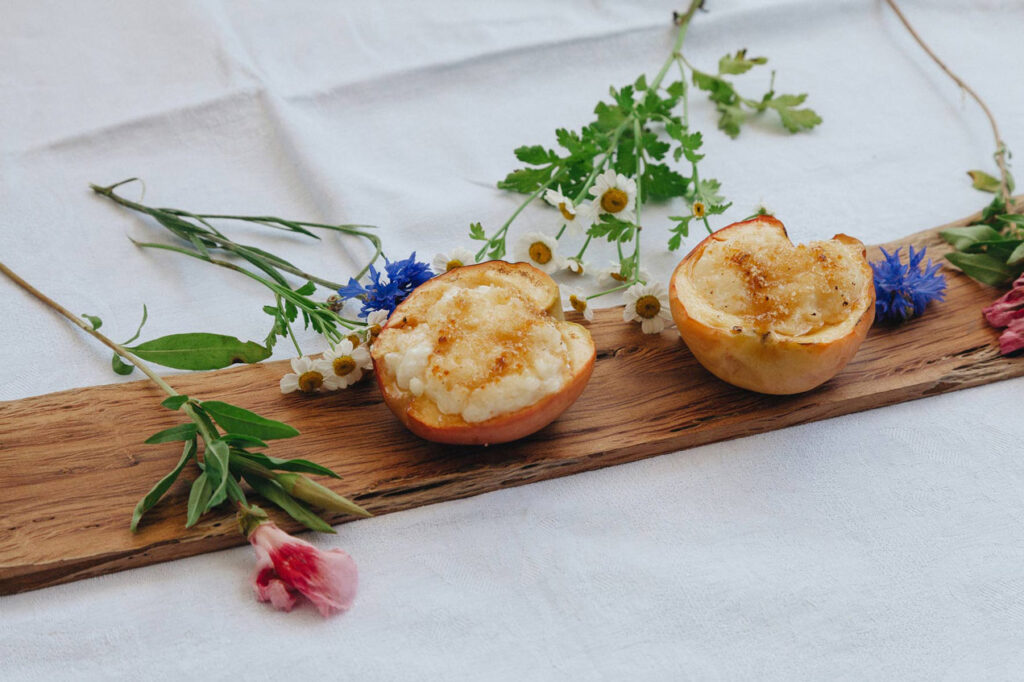 wei gebackene Äpfel mit Milchreisfüllung auf einem Holzbrett, dekoriert mit Blumen und Kräutern auf weißer Tischdecke.
