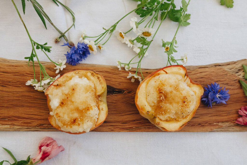 Zwei Hälften eines gebackenen Apfels, gefüllt mit cremigem Milchreis und bestreut mit Zucker und Zimt, serviert auf einem rustikalen Holzbrett, umgeben von zarten Blumen.