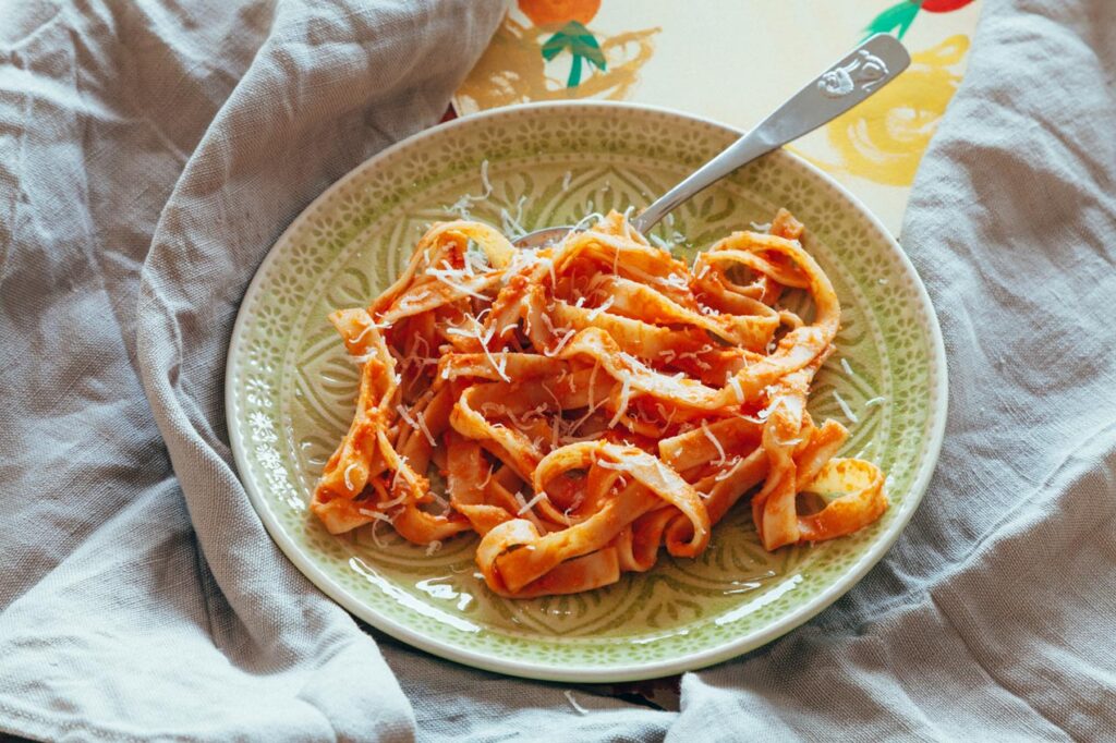 Pasta und frisch geriebenem Parmesan auf einem grünen Teller, daneben ein Besteck auf einem Leinentuch.