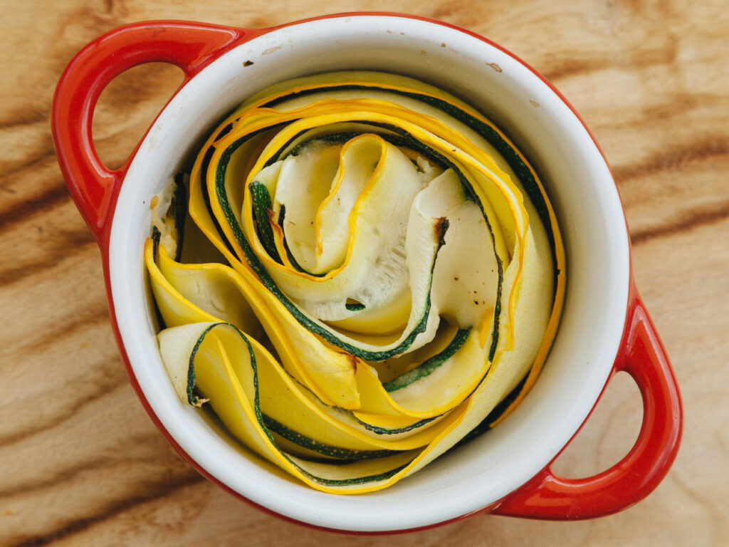 Schnelles Zucchini Rezept aus dem Ofen: Kunstvolle Zucchini-Spiralen in zwei roten Auflaufförmchen, kombiniert aus saftigen gelben und grünen Scheiben, begleitet von frischen Blüten auf natürlichem Holzuntergrund
