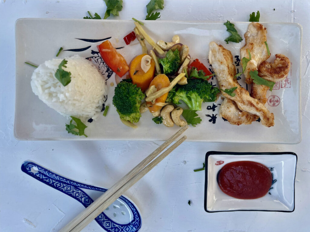Servierteller mit Gemüse, gebratenem Huhn, Reis und roter Sauce, dekoriert mit Kräutern und Essstäbchen.