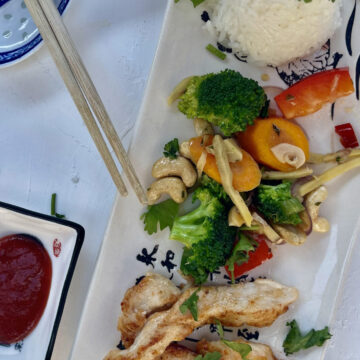 Längliche Platte mit Gemüse, gebratenem Huhn, Reisberg und rotem Dip, daneben Essstäbchen.