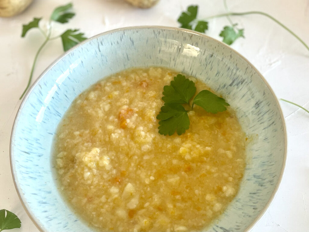 Nahaufnahme einer cremigen Kartoffelsuppe mit Möhrenstückchen und Petersilienblatt in einer blauen Schüssel.