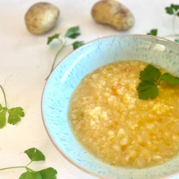 Schale mit grob pürierter, vegetarischer Kartoffelsuppe nach Omas Art, umgeben von Kartoffeln und Petersilienblättern auf weißer Fläche.
