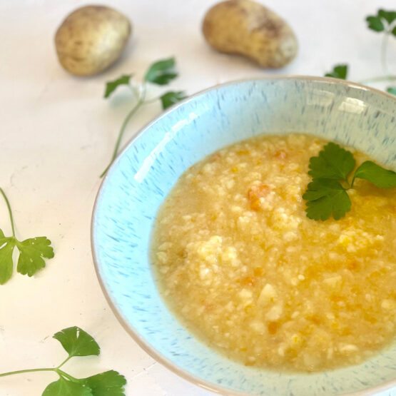 Schale mit grob pürierter, vegetarischer Kartoffelsuppe nach Omas Art, umgeben von Kartoffeln und Petersilienblättern auf weißer Fläche.