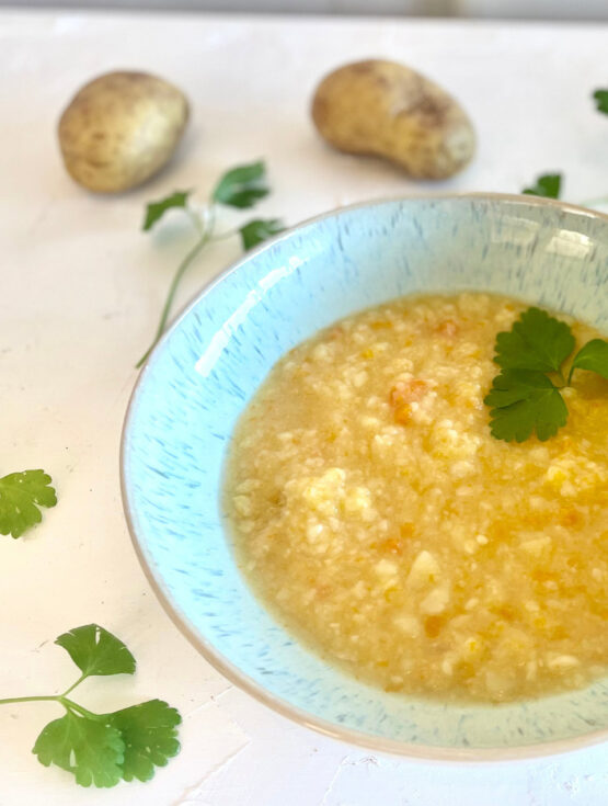 Schale mit grob pürierter, vegetarischer Kartoffelsuppe nach Omas Art, umgeben von Kartoffeln und Petersilienblättern auf weißer Fläche.