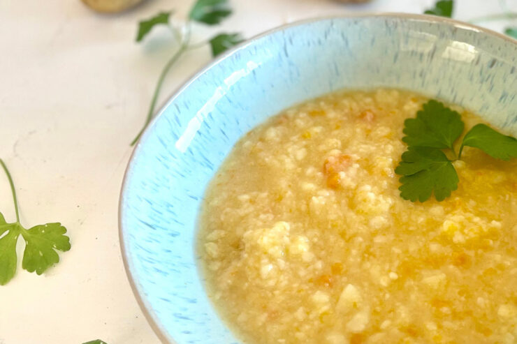 Schale mit grob pürierter, vegetarischer Kartoffelsuppe nach Omas Art, umgeben von Kartoffeln und Petersilienblättern auf weißer Fläche.